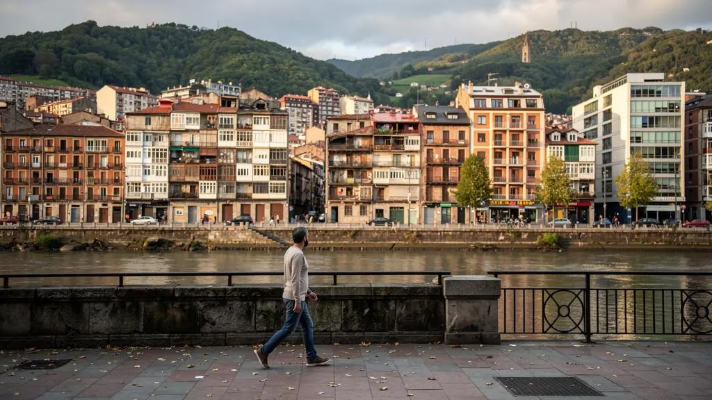 Eine Person geht am Flussufer von Bilbao entlang, im Hintergrund die kompakte Stadtlandschaft zwischen Fluss und Hügeln.