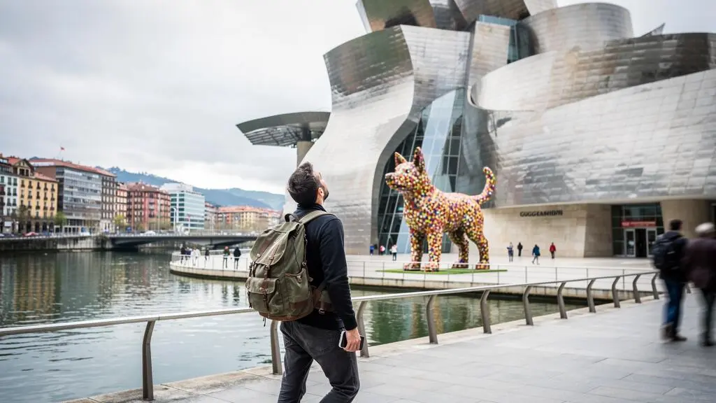 Reisende mit Rucksack stehen auf der Flusspromenade in Bilbao und blicken auf die Titanfassade des Guggenheim-Museums.
